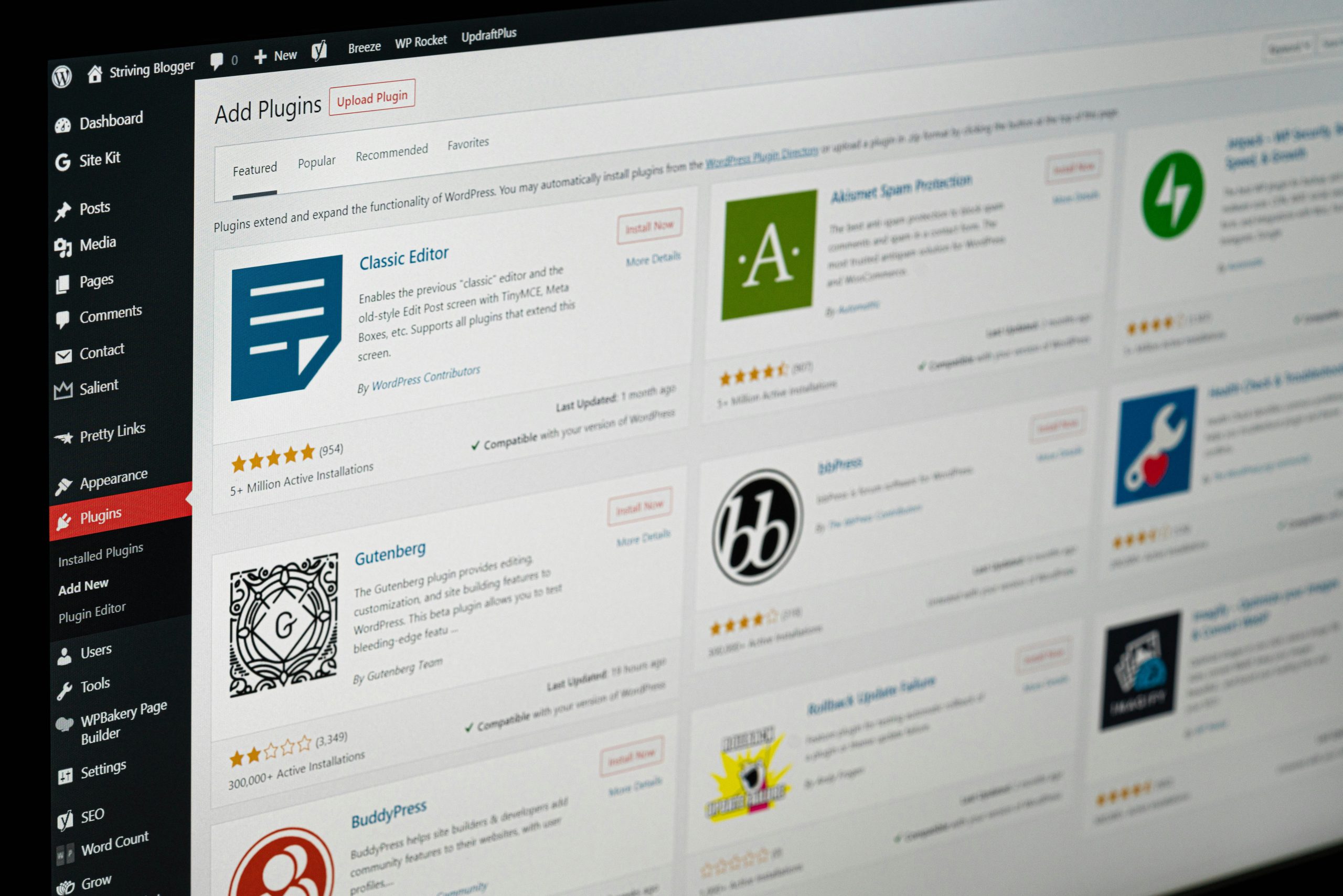 A computer screen displays the WordPress admin dashboard, showing the Add Plugins page with various plugin options, including Classic Editor, Akismet, Gutenberg, Jetpack, and BuddyPress.